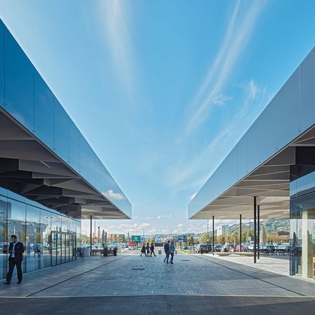 Ein Außenbereich zwischen zwei Gebäuden mit blauen Fassaden. Menschen gehen vorbei und Autos sind in der Ferne geparkt. Der Himmel ist klar mit einigen Wolken.