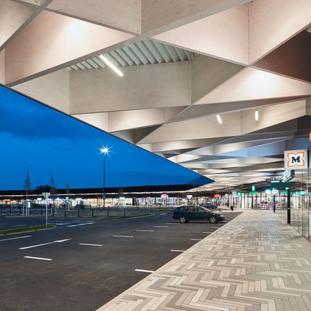 Ein Parkplatz befindet sich unter einem großen Dach, wobei der blaue Himmel sichtbar ist. Ein grünes Auto ist geparkt. Straßenlaternen und Schilder sind sichtbar.