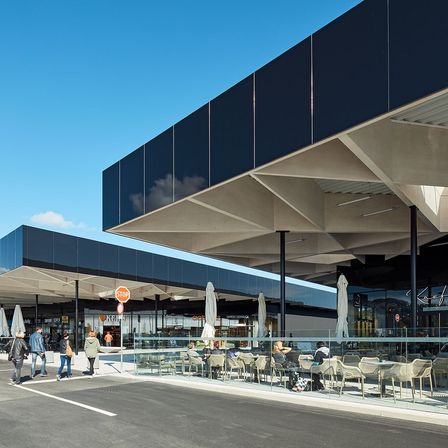 Ein modernes Gebäude mit blauen Wänden, Glaswänden und weißen Sonnenschirmen davor. Es gibt Menschen, die vor einer Glaswand gehen und auf Stühlen sitzen. Links befindet sich ein Stoppschild.