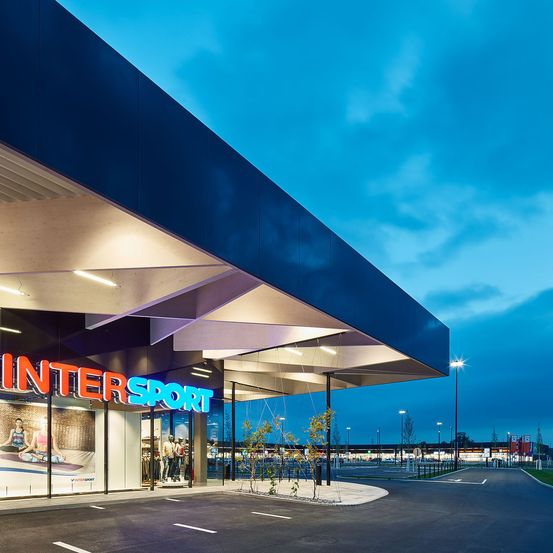 Der Eingang eines Intersport-Geschäfts wird bei Dämmerung von seinem roten Neonschild beleuchtet, mit einem Parkplatz im Vordergrund.