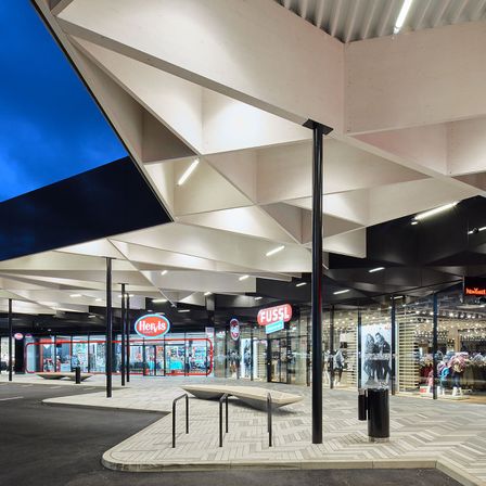 Ein überdachtes Einkaufszentrum mit geometrischem Deckendesign. Zwei Geschäfte sind sichtbar, eines heißt Hervis Sports und das andere Fussl. Der Boden hat ein Muster aus Ziegeln.