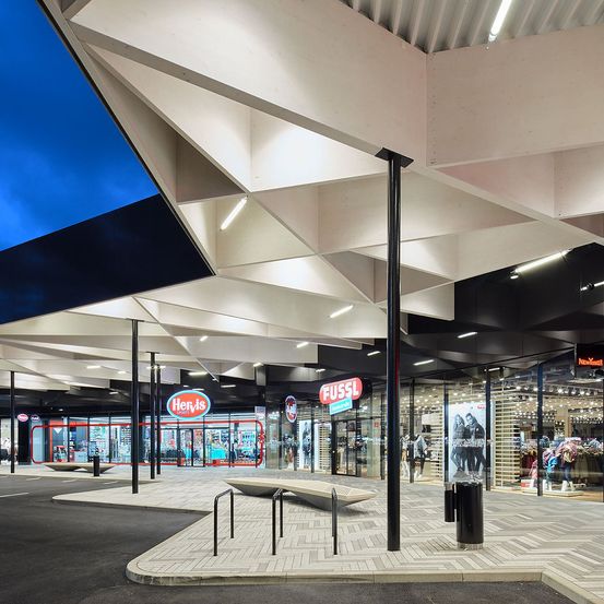Ein überdachtes Einkaufszentrum mit geometrischem Deckendesign. Zwei Geschäfte sind sichtbar, eines heißt Hervis Sports und das andere Fussl. Der Boden hat ein Muster aus Ziegeln.