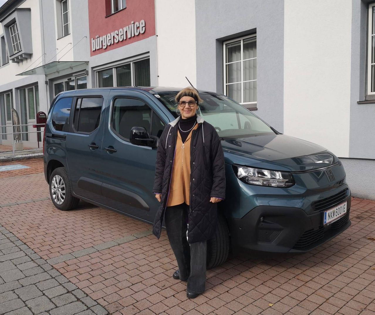 A woman stands next to a dark blue van with a license plate, in front of a building with a sign reading Burgerservice.