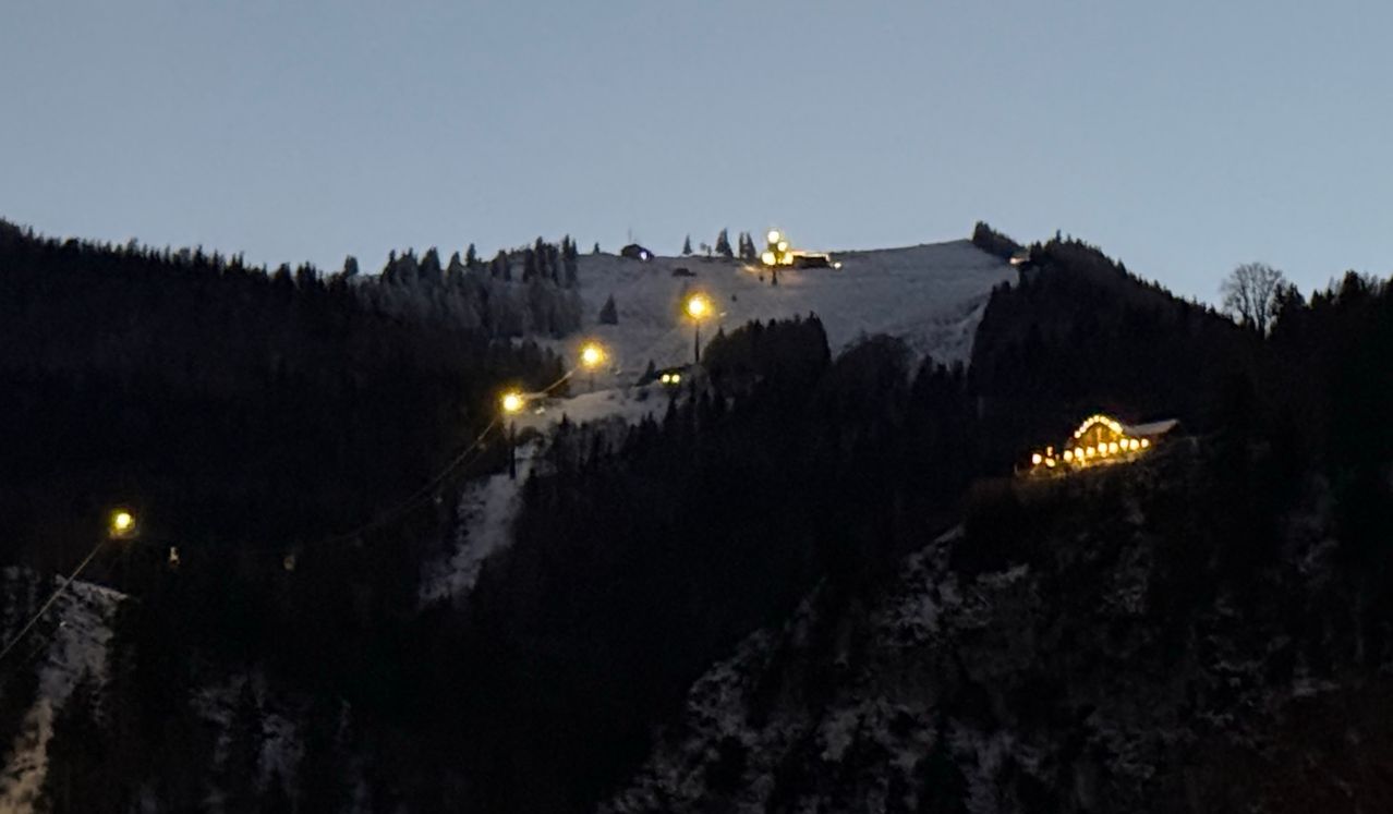 Ein verschneiter Berg mit Kiefern bei Dämmerung, beleuchtet von mehreren gelben Lichtern am Hang und einem hellen Licht in der Ferne.