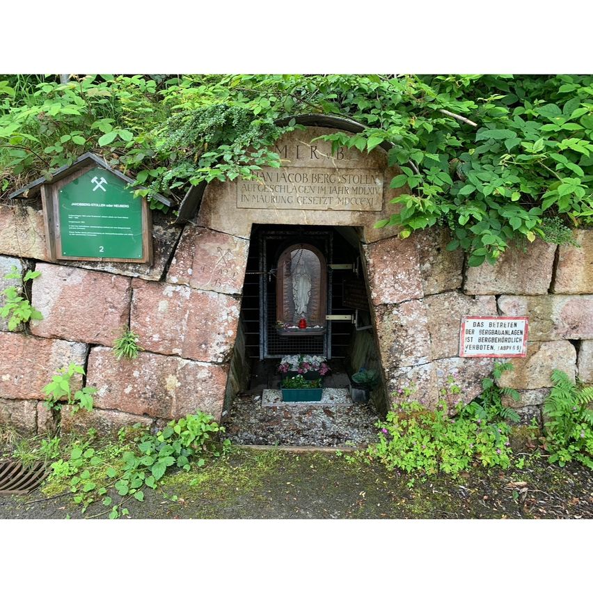Ein steinerner Eingang mit einer Statue eines Heiligen im Inneren. Es ist von Grün umgeben und hat ein Schild an der Wand mit lateinischem Text.