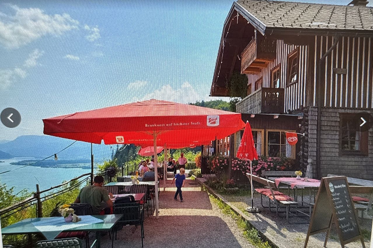 Ein Outdoor-Restaurant mit Blick auf einen See. Ein Kind geht unter einem großen roten Schirm. Tische und Stühle sind aufgestellt, und es gibt ein Gebäude mit Balkon.
