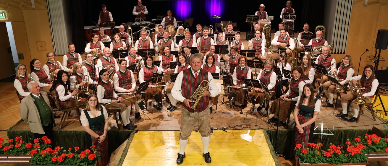 Eine Band in traditioneller Kleidung spielt auf der Bühne mit Musikinstrumenten, während das Publikum dahinter sitzt und zwei Frauen auf beiden Seiten der Bühne stehen.