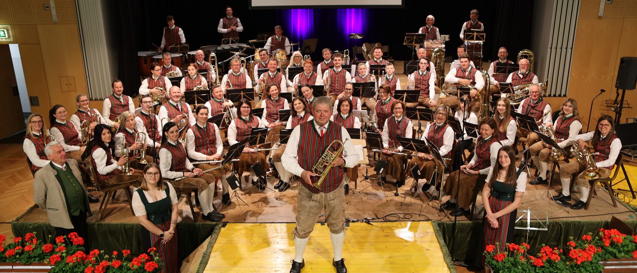 Eine Band in traditioneller Kleidung spielt auf der Bühne mit Musikinstrumenten, während das Publikum dahinter sitzt und zwei Frauen auf beiden Seiten der Bühne stehen.