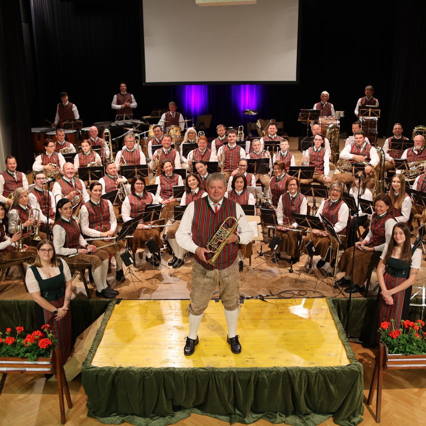 Eine Band in traditioneller Kleidung spielt auf der Bühne mit Musikinstrumenten, während das Publikum dahinter sitzt und zwei Frauen auf beiden Seiten der Bühne stehen.