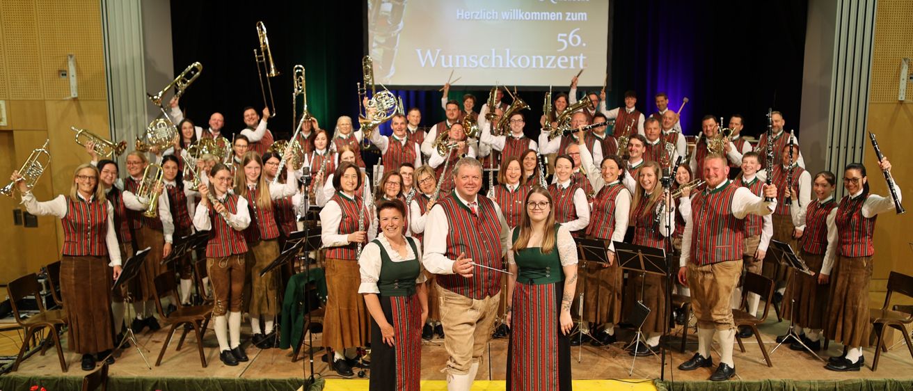 Ein Blasorchester posiert für ein Foto auf der Bühne mit einer großen Leinwand im Hintergrund. Die Leinwand zeigt 'Herzlich willkommen zum 56. Wunschkonzert'. Die Bandmitglieder sind in passenden Outfits gekleidet.