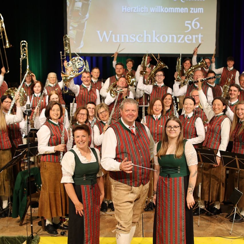 Ein Blasorchester posiert für ein Foto auf der Bühne mit einer großen Leinwand im Hintergrund. Die Leinwand zeigt 'Herzlich willkommen zum 56. Wunschkonzert'. Die Bandmitglieder sind in passenden Outfits gekleidet.