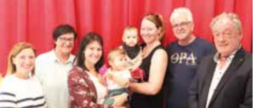 Eine Gruppe von Menschen steht lächelnd vor einem roten Vorhang, um ein Foto zu machen. Zwei Frauen halten ein Baby, während ein Mann in der Mitte ein blaues T-Shirt trägt.