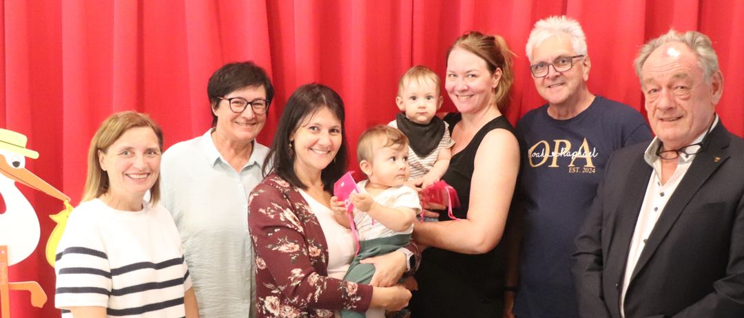 Eine Gruppe von Menschen, die vor einem roten Vorhang stehen, lächeln und für ein Foto posieren. Eine Frau hält ein Baby in den Armen.