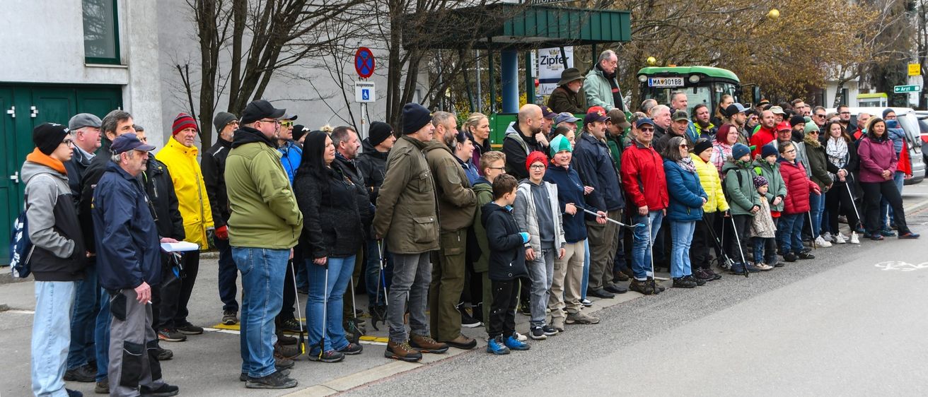 Eine Gruppe von Menschen mit Stöcken und Mützen steht auf einer Straße, mit einem Traktor und einem Informationsschild im Hintergrund.