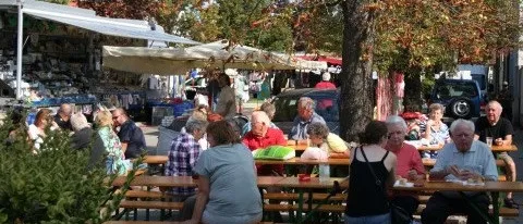 Eine Gruppe von Menschen sitzt an Tischen auf einem Freiluftmarkt und genießt Essen und Getränke unter einem Baldachin.
