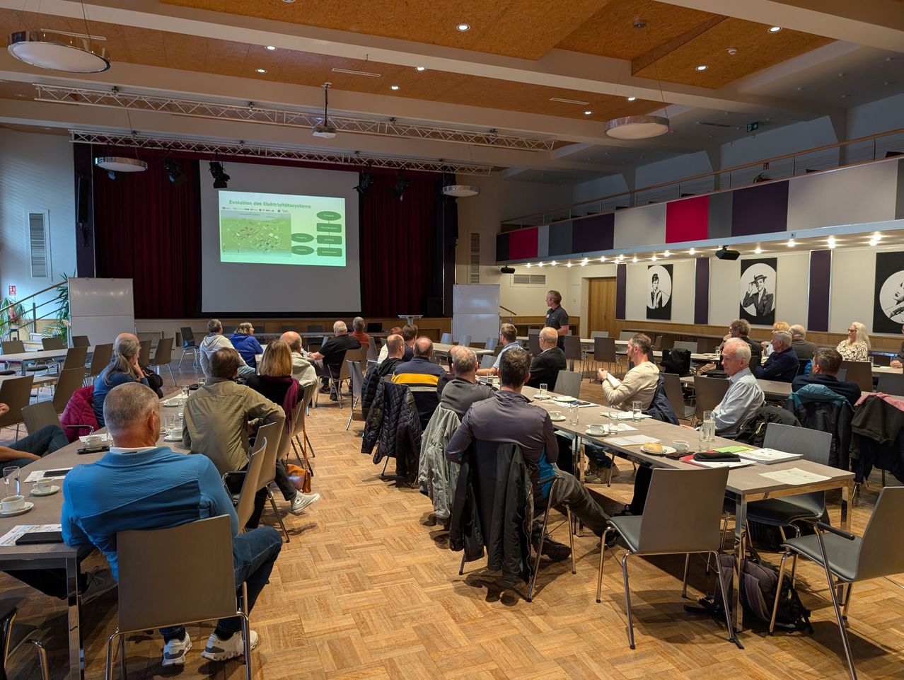 Eine Gruppe von Menschen sitzt in einem Konferenzsaal an Tischen, vor einer Leinwand mit einer Folie. Ein Mann steht vorne und präsentiert möglicherweise. Der Raum hat einen Holzboden, einen roten Vorhang und Porträts an der Wand.