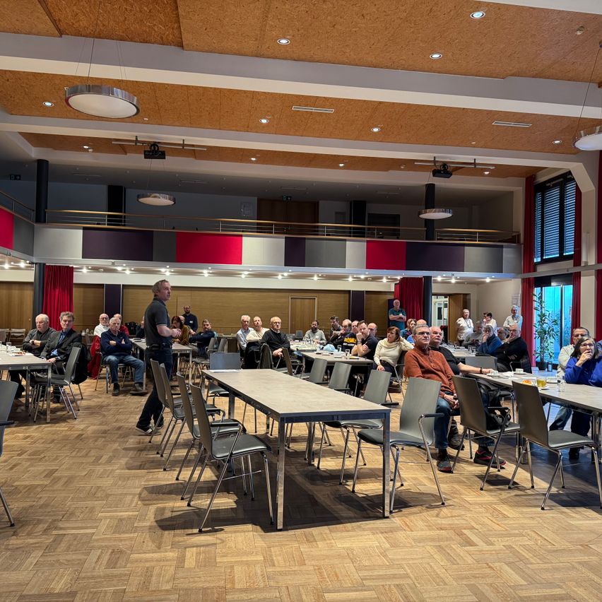 Ein großer Raum mit Holzboden, roten Vorhängen und Tischen, die für eine Versammlung vorbereitet sind. Ein Mann steht vor einer Gruppe von Menschen.