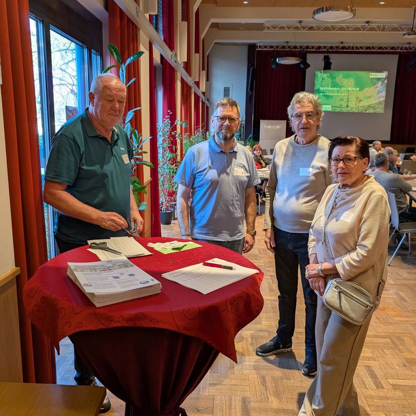Vier Erwachsene stehen um einen Tisch mit Papieren und Scheren. Der Tisch ist mit rotem Stoff bedeckt. Eine Frau trägt eine Handtasche. Eine Projektionsleinwand befindet sich hinter ihnen.