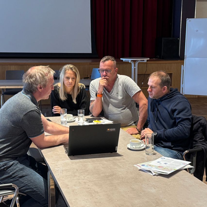 Vier Personen sitzen in einem Konferenzraum um einen Tisch herum, vor einer Projektionsleinwand. Ein Laptop ist offen, und es gibt Gläser und Papiere auf dem Tisch.