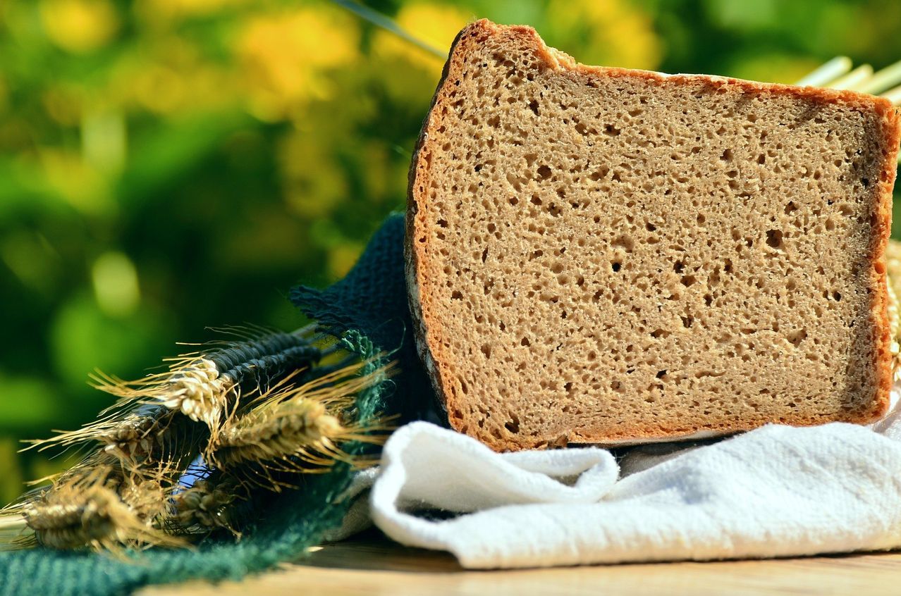 A slice of rustic bread with a few holes sits beside a towel and some wheat on a wooden table.