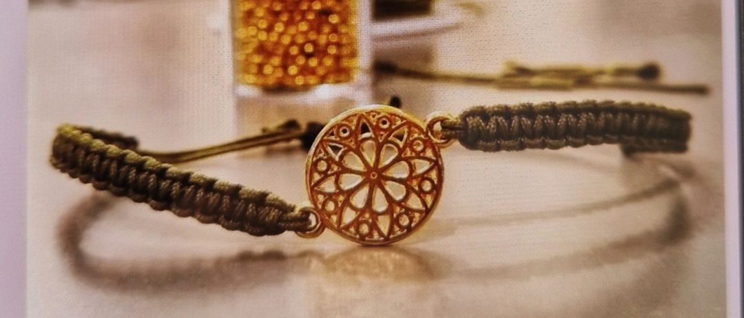 Ein Armband mit goldenem Zentrum und braunem Seil liegt auf einem Tisch mit verschwommenem Hintergrund. Das Bild hat einen Wasserzeichen mit den Worten 'Kreativ Workshops' in weiß.