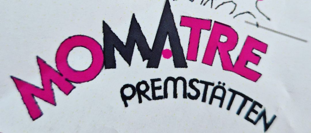 Das 'Momatre Premium'-Etikett ist auf einem weißen Hintergrund mit rosa und schwarzen Farben gedruckt. Das Etikett hat eine Skizze einer rennenden Familie und der Text ist in einer stilisierten Schrift.