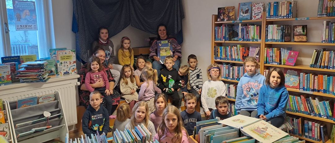 Eine Gruppe von Kindern und Erwachsenen versammelt sich in einer Bibliothek, wobei ein Erwachsener vorne sitzt und ein Buch hält. Kinder sitzen und stehen vor den Bücherregalen.