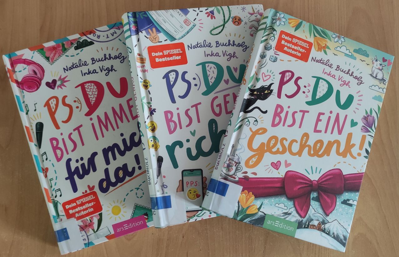 Three books with colorful covers are displayed on a wooden surface. The first book has a cat and a ribbon on its cover. The second book has a colorful design with the word 'PS' in green. The third book has a black cat on its cover.