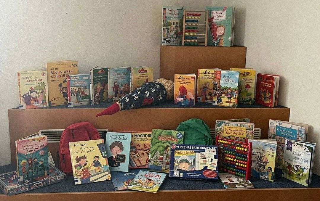 A display of children's books, toys, and educational materials on a wooden shelf. A red backpack and an abacus are also visible.