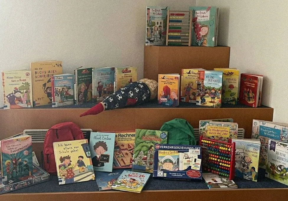 Eine Ausstellung von Kinderbüchern, Spielzeug und Lernmaterialien auf einem Holzregal. Ein roter Rucksack und eine Rechenkette sind ebenfalls sichtbar.