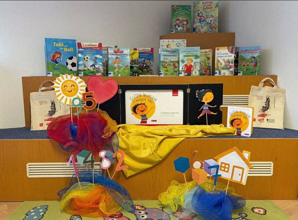 Ein Bücherregal mit Kinderbüchern, darunter 'Tobi und der Ball' und 'Leseloewen', ist mit bunten Dekorationen und Zahlen verziert. Die Bücher sind von anderen Spielzeugen und Dekorationen umgeben.