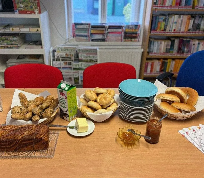 Ein Tisch mit Brot, Brötchen und Marmelade. Eine Milchtüte, Butter und ein Stapel Teller. Zwei rote Stühle und Bücherregale im Hintergrund.