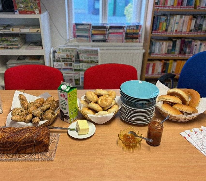 Ein Tisch mit Brot, Brötchen und Marmelade. Eine Milchtüte, Butter und ein Stapel Teller. Zwei rote Stühle und Bücherregale im Hintergrund.