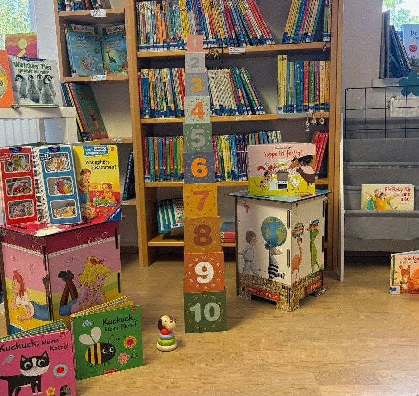 Eine Kinderbuchecke mit einem hölzernen Bücherregal gefüllt mit Büchern. Ein bunter Spielzeugturm mit Zahlen steht auf dem Boden, neben einem kleinen Spielzeug und einem Buch mit einer Katze.
