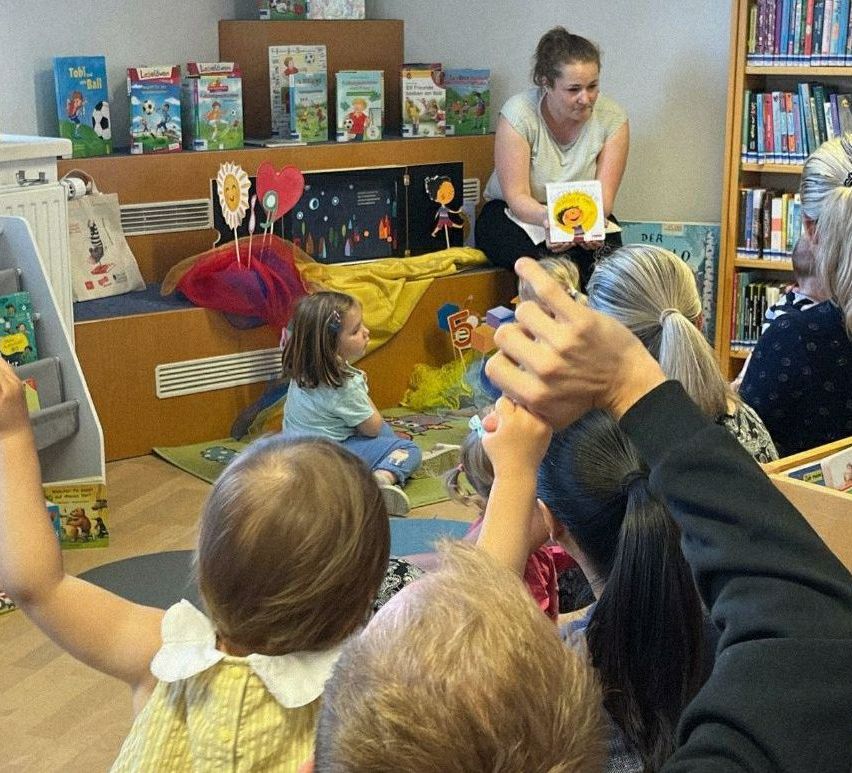 Eine Frau liest Kindern in einem Raum mit Bücherregalen, Spielzeug und einem farbenfrohen Hintergrund vor. Die Kinder sitzen auf dem Boden und lauschen aufmerksam.