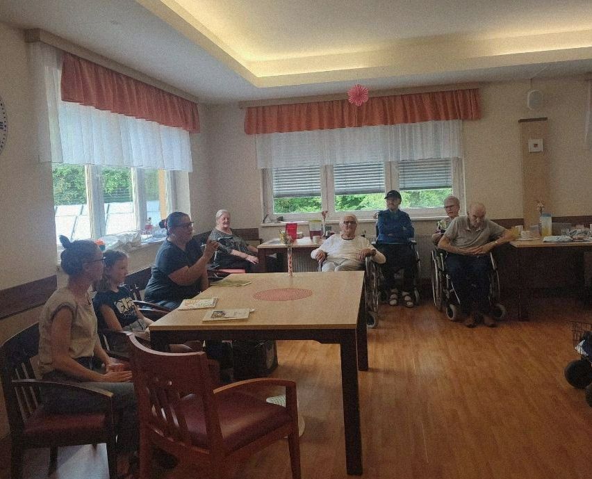 Eine Gruppe von Menschen sitzt in einem Raum, wahrscheinlich einem Pflegeheim. Einige sind im Rollstuhl, andere sitzen in Stühlen. Eine Tisch steht in der Mitte und eine Frau spricht in ein Mikrofon. Der Raum hat Fenster mit Vorhängen und eine dekorative Blume an der Decke.