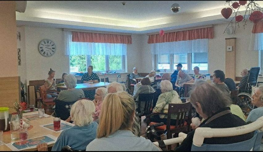 In einem Raum mit einer Uhr an der Wand sitzen ältere Menschen in Stühlen, einige in Rollstühlen. Sie diskutieren wahrscheinlich etwas, während eine Person vor dem Tisch steht.
