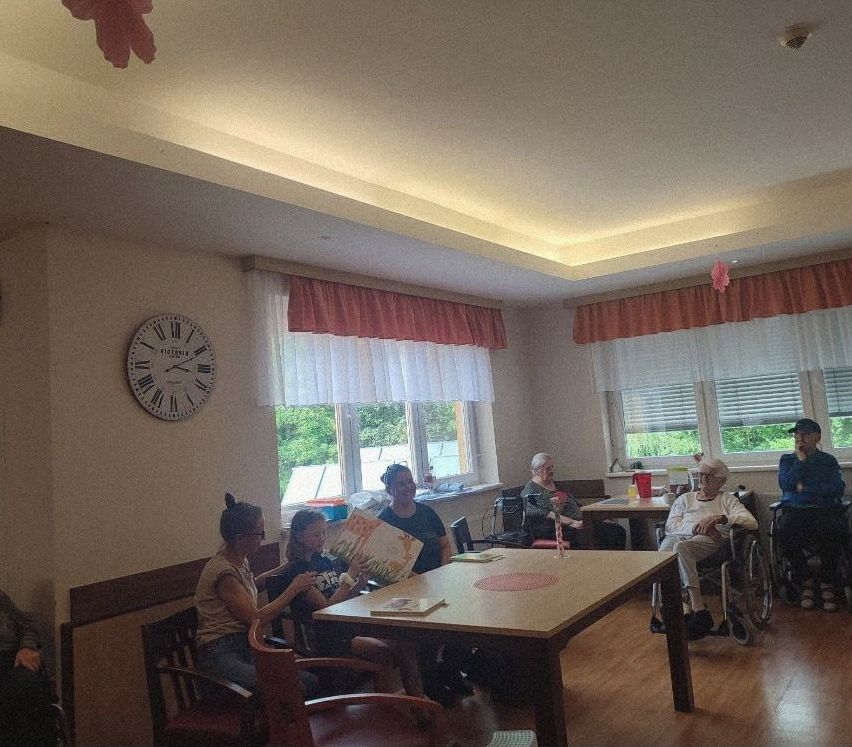 Eine Gruppe von Menschen, darunter Frauen und ein älterer Mann im Rollstuhl, sitzt um einen Tisch in einem Raum. Eine Uhr ist an der Wand.