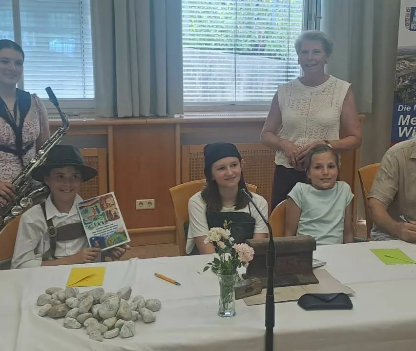 Eine Gruppe von Kindern sitzt an einem Tisch mit Steinen, Blumen und einem Mikrofon. Sie lächeln für ein Foto. Eine Frau steht hinter ihnen und lächelt.