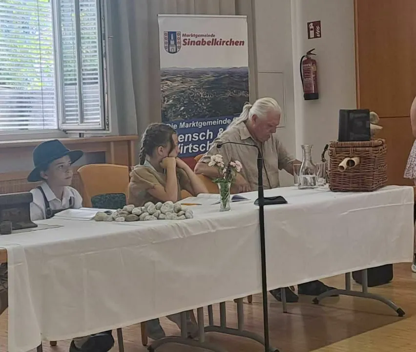 Ein Mann und zwei Kinder sitzen an einem Tisch. Der Mann spricht, und die Kinder hören aufmerksam zu. Ein Mikrofon steht vor dem Mann, und ein Korb ist auf dem Tisch.