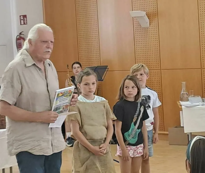 Ein älterer Mann hält ein Buch und spricht mit zwei Mädchen und einem Jungen in einem Raum mit einer Holzwand und einem Feuerlöscher.