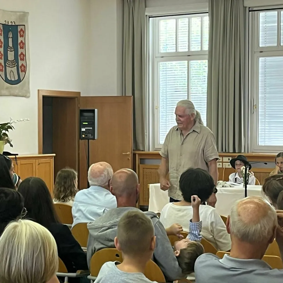 Eine Indoor-Versammlung mit einem Redner vor einem sitzenden Publikum, ein erwachsener Mann mit grauem Haar, der spricht, während andere aufmerksam zuhören, eine dekorierte Wand und ein Topfpflanzen.