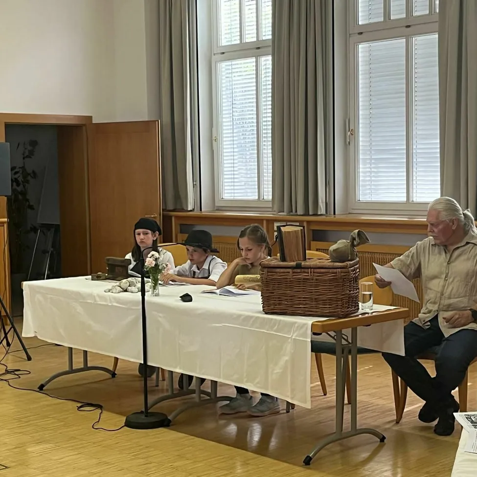 Eine Gruppe von Kindern und ein älterer Mann sitzen um einen Tisch herum. Der Mann liest ein Papier, während die Kinder ihre Bücher studieren. Ein Korb und Blumen stehen auf dem Tisch.