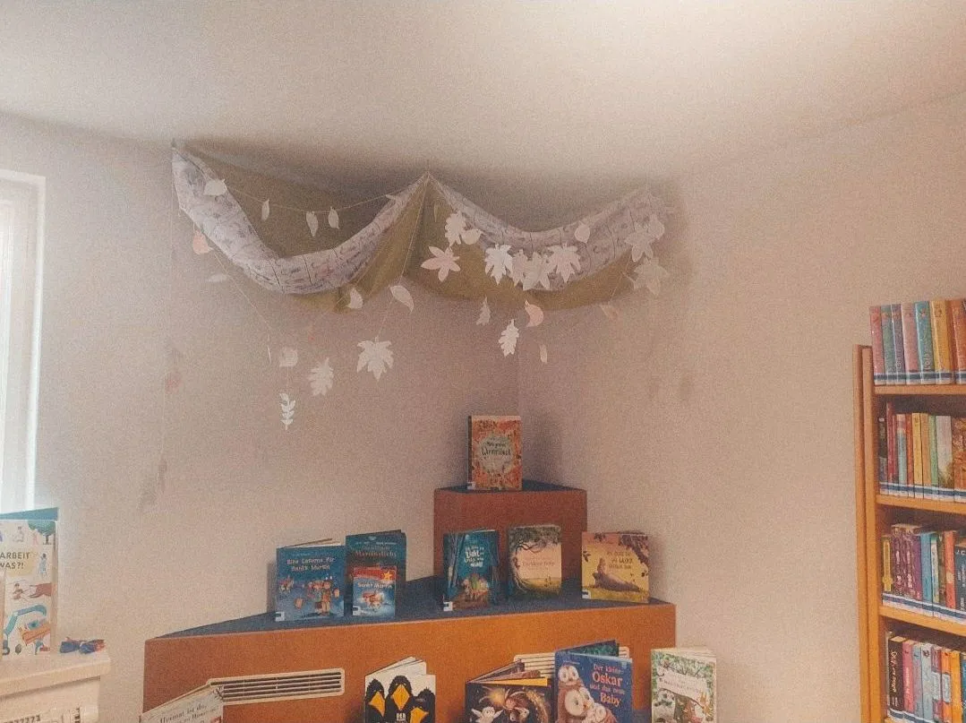 Eine gemütliche Ecke in einem Raum mit weißer Wand und Decke. Ein Regal mit Büchern und eine dekorative Girlande aus Blättern, die von der Decke hängen.