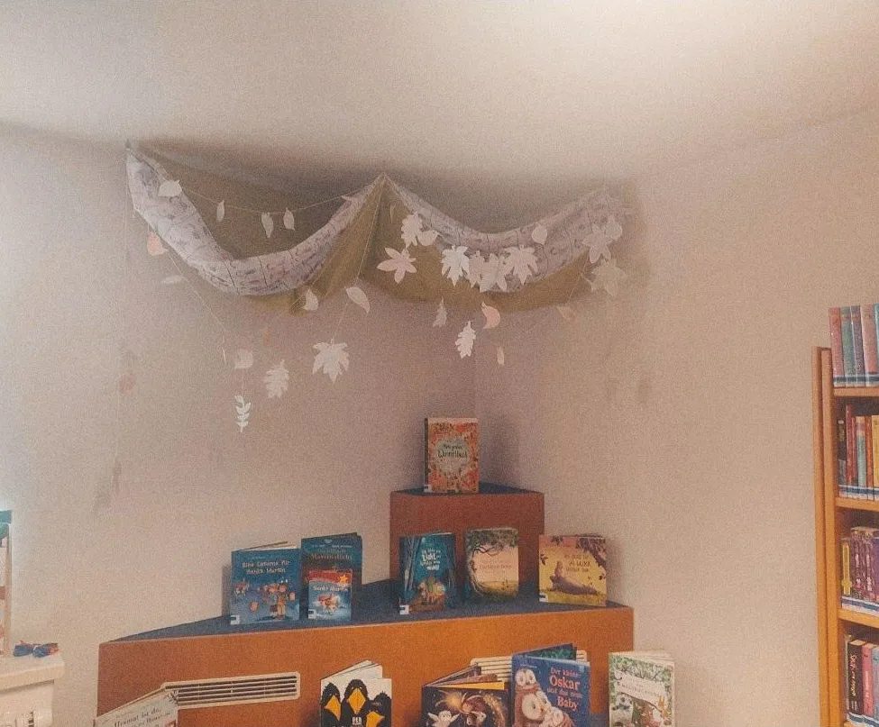 Eine gemütliche Ecke in einem Raum mit weißer Wand und Decke. Ein Regal mit Büchern und eine dekorative Girlande aus Blättern, die von der Decke hängen.