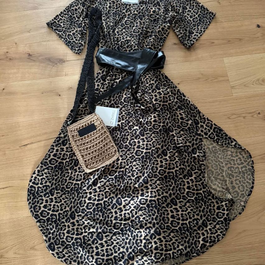 Ein Leopardenmuster-Kleid mit einem schwarzen Gürtel und einer kleinen gewebten Tasche liegt auf einem Holzboden.
