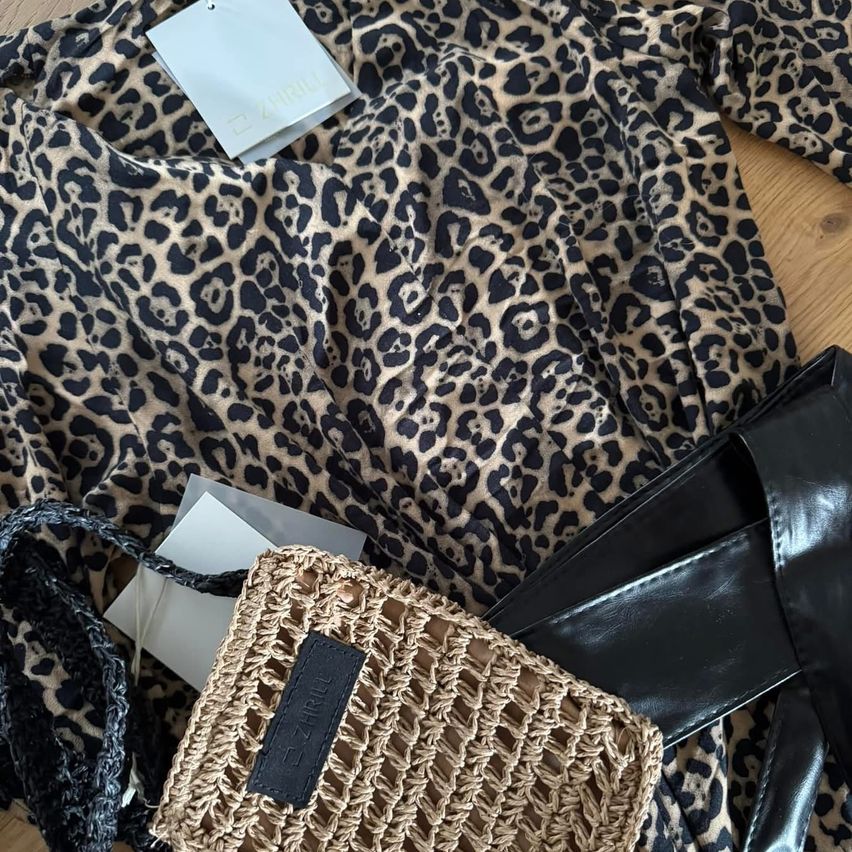 Ein Leopardenprint-Bluse mit einem Etikett darauf liegt auf einem Tisch. Daneben befindet sich eine gehäkelte Tasche und ein schwarzes Lederaccessoire.