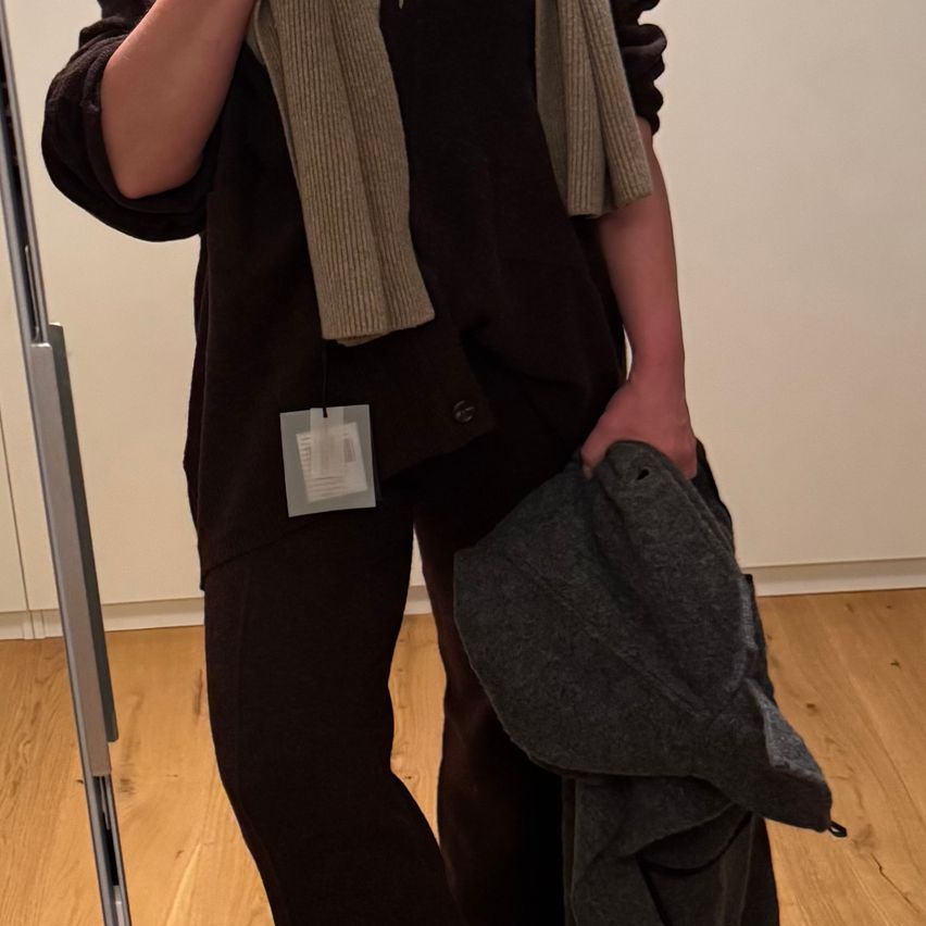 Eine Person steht in einem Raum mit Holzboden, hält einen grauen Mantel und trägt ein schwarzes Oberteil und eine Schürze. Es gibt einen weißen Aufkleber am Oberteil.