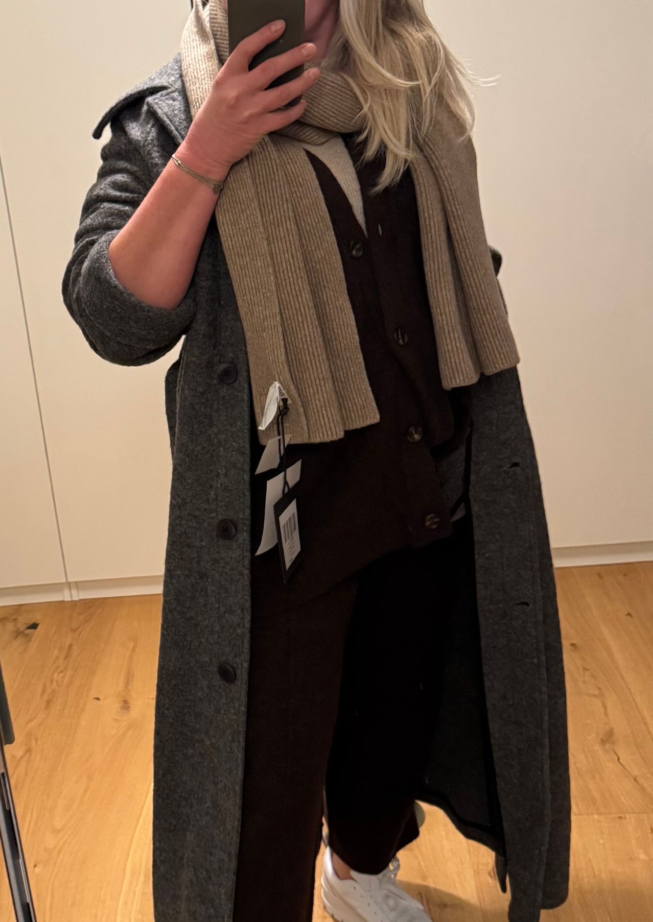 Eine Frau in einem grauen Mantel und einem Schal um den Hals steht in einem Raum mit Holzböden. Sie hat ihre Hand am Kinn und ein Etikett ist an ihrem Mantel sichtbar.