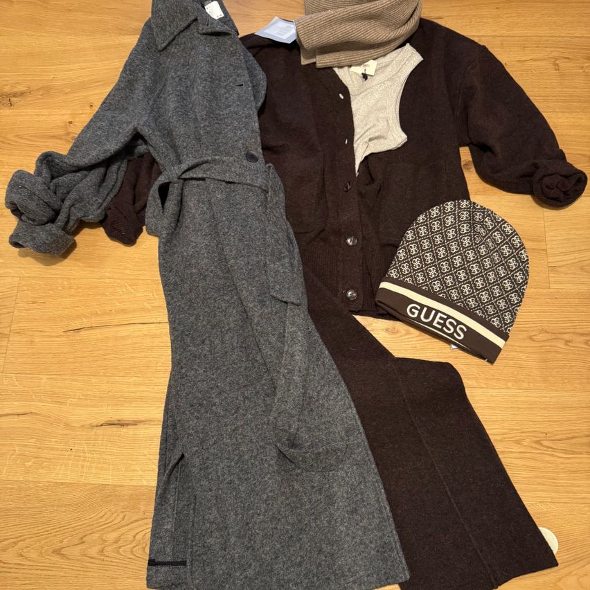 Eine Auswahl an Kleidungsstücken auf einem Holzboden, darunter ein grauer Trenchcoat, ein brauner Strickpullover, eine braune und weiße Mütze und ein brauner Schal.